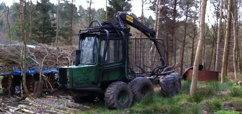 Green forestry forwarder with a loading crane parked in pine woodland beside a stack of cut timber